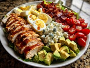 A beautifully arranged plate featuring a classic Cobb salad with sliced grilled chicken, bacon, hard-boiled eggs, tomatoes, avocado, and blue cheese.