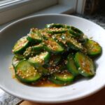 Close-up of sliced cucumber salad coated in a savory, orange-brown dressing and topped generously with toasted sesame seeds.