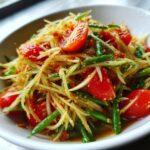 Close-up of vibrant papaya salad featuring shredded green papaya, cherry tomatoes, and green beans in a savory dressing.