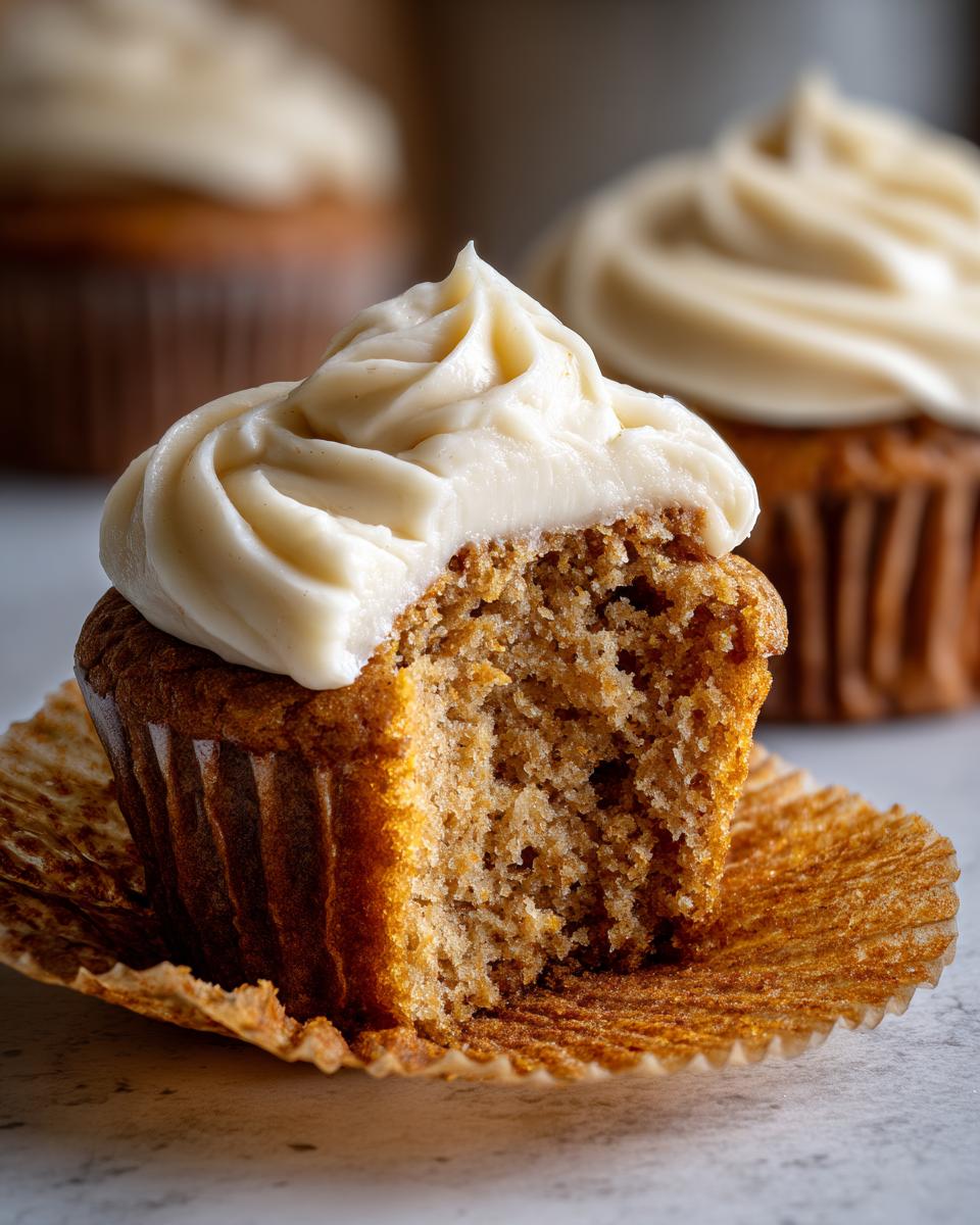 A close-up of a banana cupcakes with a bite taken out, showing the moist crumb and thick cream cheese frosting.