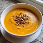 A close-up of vibrant orange carrot ginger soup garnished with toasted pumpkin seeds.