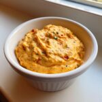 A close-up of thick, orange-yellow cottage cheese queso dip speckled with red peppers and green herbs in a white ramekin.