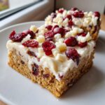 A close-up of a square of cranberry bliss bars topped with white frosting, dried cranberries, and white chocolate chunks.