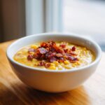 A creamy bowl of corn chowder topped generously with crispy bacon bits, sitting on a wooden table.