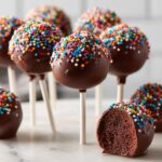 Close-up of several chocolate cake pops covered in rainbow sprinkles, one is cut open showing the rich interior.