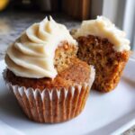 Two banana cupcakes on a white plate, one with a bite taken out, showing moist crumb and cream cheese frosting.