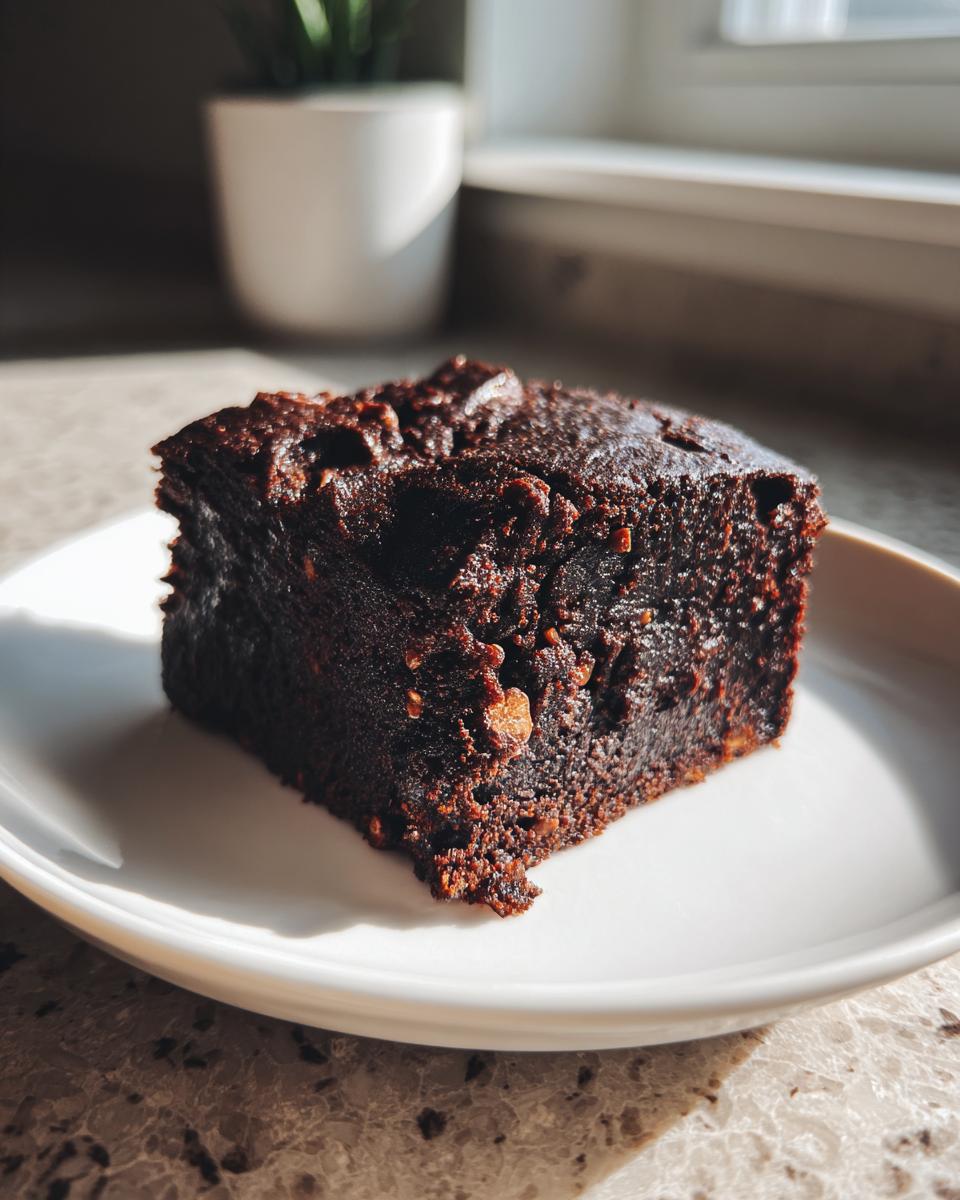 A rich, fudgy square of date brownies sits on a white plate, bathed in sunlight.