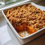 A square white baking dish filled with golden brown pumpkin crisp, showing a serving removed.