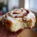 A close-up of a gooey cinnamon roll being held, showing a bite taken out of the soft interior and thick cream cheese frosting.