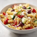 A close-up shot of a white bowl filled with vibrant greek pasta salad featuring rotini pasta, cucumbers, tomatoes, olives, and feta.
