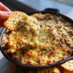 A hand scoops cheesy, hot crab dip recipe from a cast iron skillet using a round cracker.