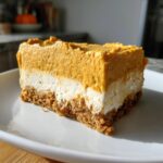 A thick, layered slice of pumpkin cheesecake bar with a graham cracker crust, cream cheese layer, and pumpkin topping.