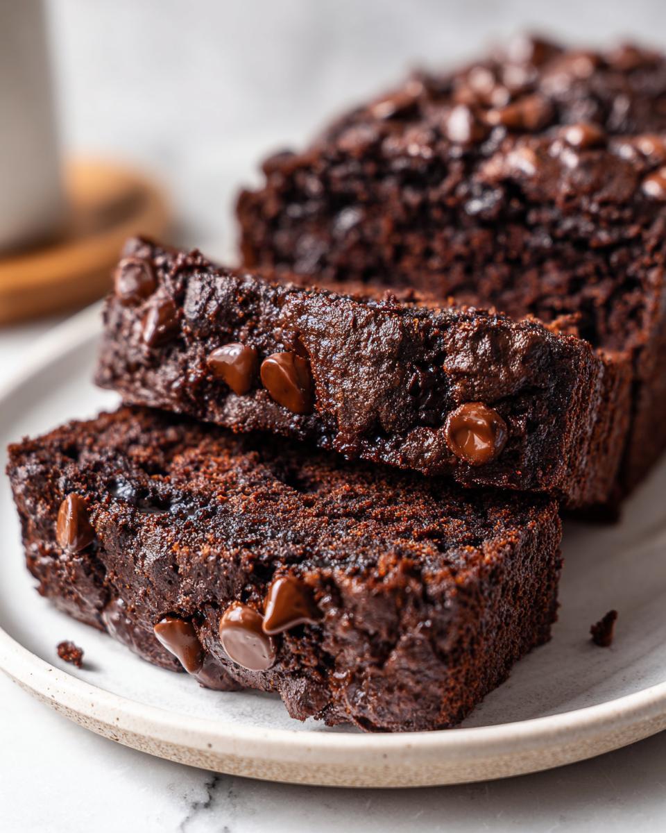 Close-up of two moist slices of rich chocolate zucchini bread topped with melted chocolate chips on a white plate.