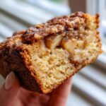 Close-up of a moist slice of jewish apple cake held by a hand, showing tender cake crumb and cinnamon-spiced apple chunks.