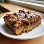 Close-up of a rich, layered cookie bars slice topped with caramel, chocolate chips, and sea salt.