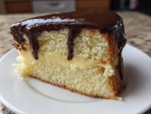 A close-up of a slice of Boston Cream Pie featuring vanilla cake, pastry cream filling, and dripping chocolate ganache.