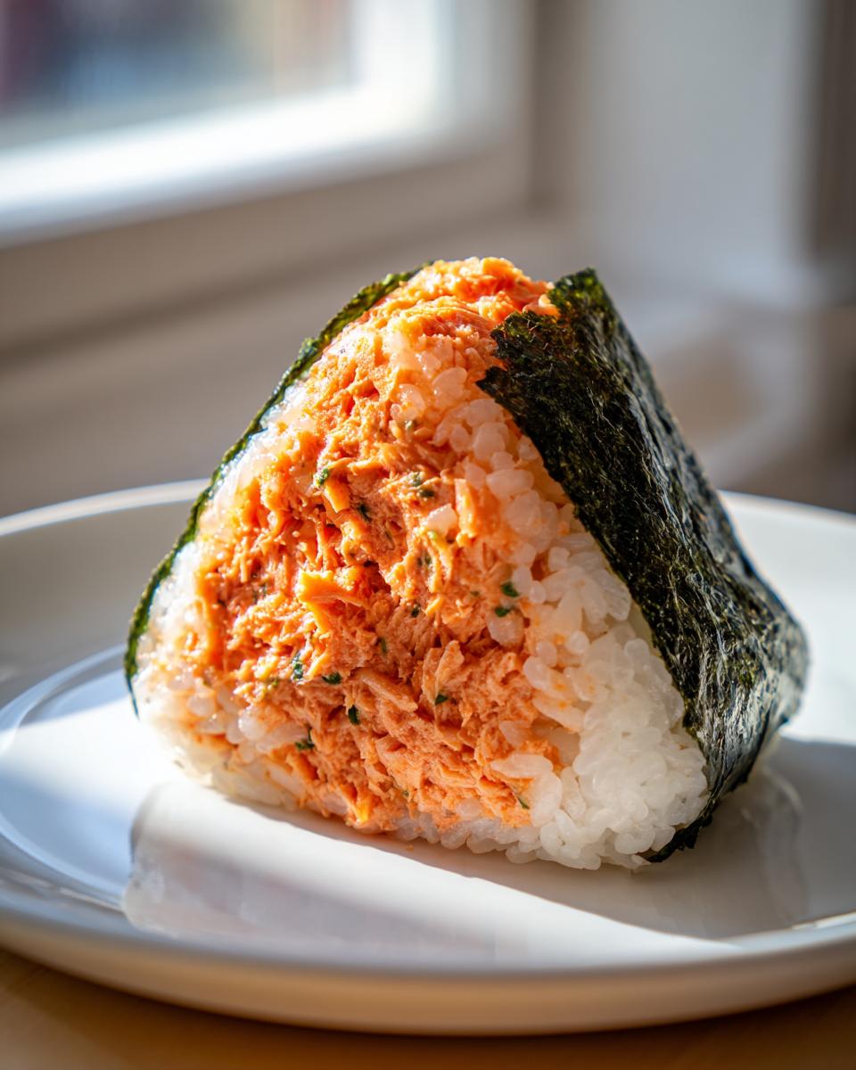 A triangular tuna onigiri filled with bright orange spicy tuna mixture, wrapped partially in dark nori seaweed, resting on a white plate.