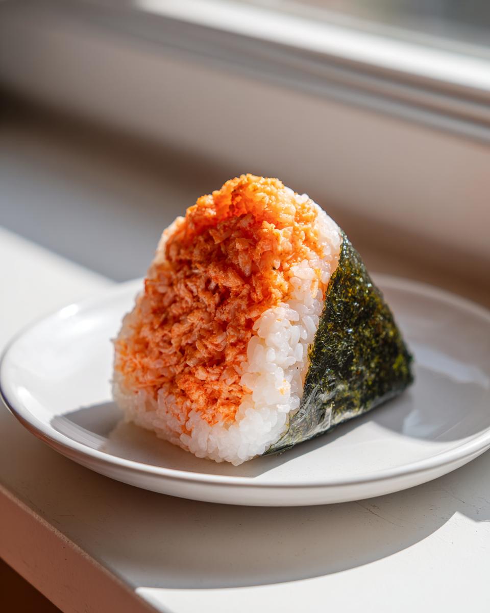 A single triangular tuna onigiri, topped with spicy orange seasoning, resting on a white plate near a window.