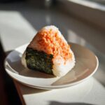 A single, triangular tuna onigiri resting on a white plate, partially wrapped in dark nori seaweed.