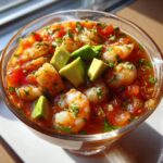 A close-up, vibrant image of mexican shrimp cocktail served in a clear glass bowl, topped with avocado chunks and cilantro.