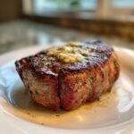 Close-up of a perfectly seared air fryer steak topped with melting garlic herb butter on a white plate.