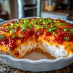 A serving removed from a baked cream cheese dip topped with melted cheese, red sauce, and green onions.