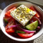 A vibrant, close-up shot of a traditional greek salad featuring a large block of feta cheese drizzled with olive oil and herbs over tomatoes, cucumbers, and red onion.