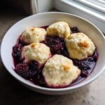 A white bowl filled with warm Blackberry Dumplings swimming in a rich, dark purple blackberry sauce.