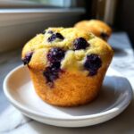 A close-up of a golden baked blueberry cottage cheese muffin sitting on a small white saucer.