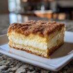 A thick, layered slice of churro cheesecake bars featuring a crust, creamy filling, and cinnamon sugar topping.
