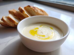 A white bowl of creamy garlic soup topped with olive oil and pepper, served with sliced baguette.