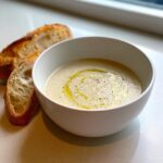 A white bowl filled with creamy garlic soup, drizzled with olive oil and black pepper, served with crusty bread.