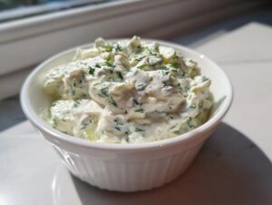 A close-up of a small white bowl filled with thick, creamy homemade tartar sauce recipe, garnished with fresh green herbs.