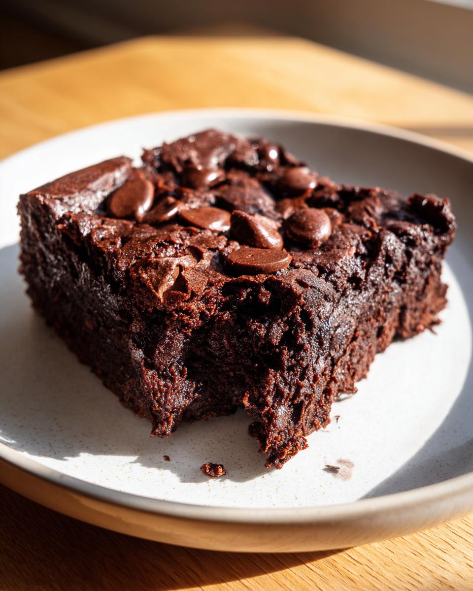 Close-up of a rich, dark slice of black bean brownies topped with melted chocolate chips on a speckled white plate.