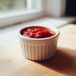 A small white ramekin filled with chunky red homemade cocktail sauce recipe, sitting on a wooden surface near a bright window.