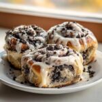 Three decadent oreo cinnamon rolls covered in white icing and crushed dark cookie pieces, served on a white plate.