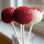 Three vibrant red velvet cake pops, two fully coated in red crumbs and one dipped in white chocolate, displayed in a small glass.