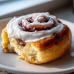 A close-up, appetizing shot of a single sourdough discard cinnamon roll topped with thick white icing, sitting on a white plate.