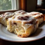 Close-up of four fluffy tiktok cinnamon rolls covered in thick white icing on a white plate near a window.